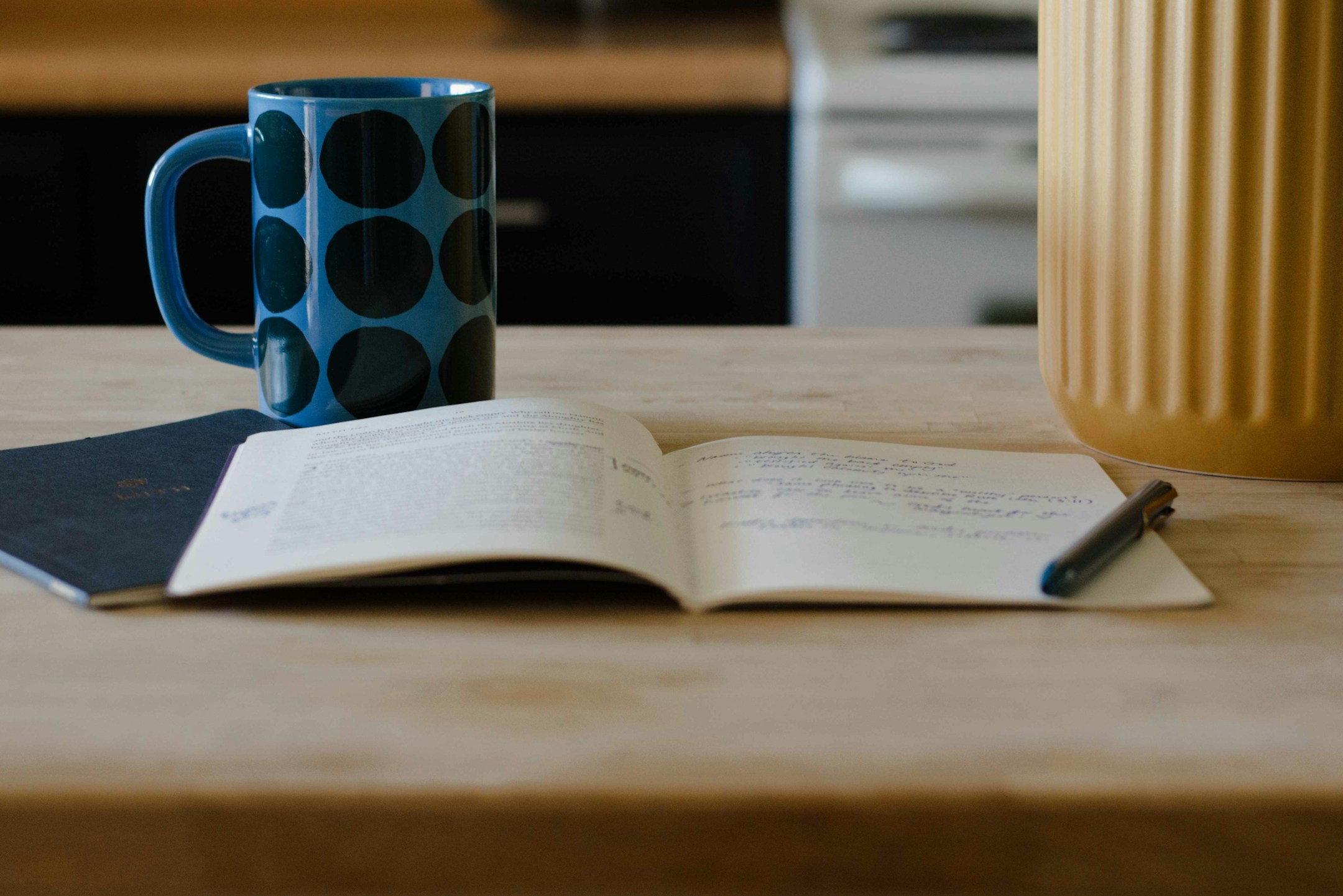 A ceramic mug, a notebook, a pen on a wooden surface, soft ambient warmth, the still moment of writing something down.