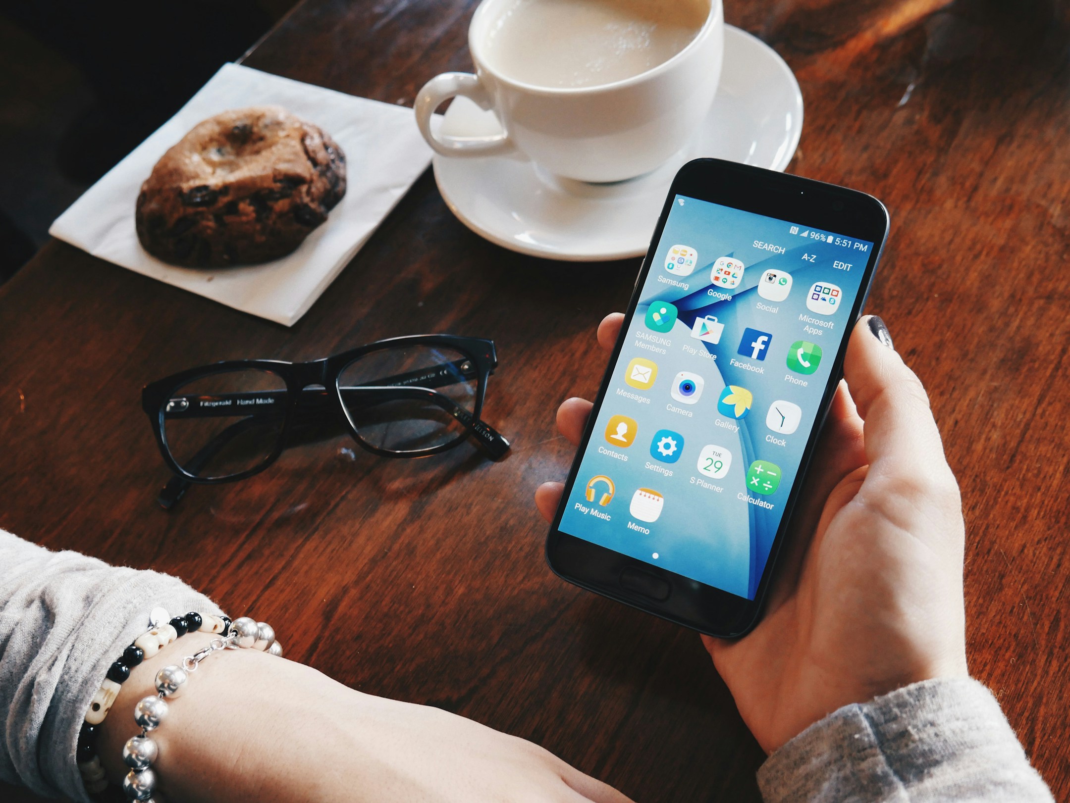 A hand holding a phone over a ceramic cup on a wooden table, soft morning light.
