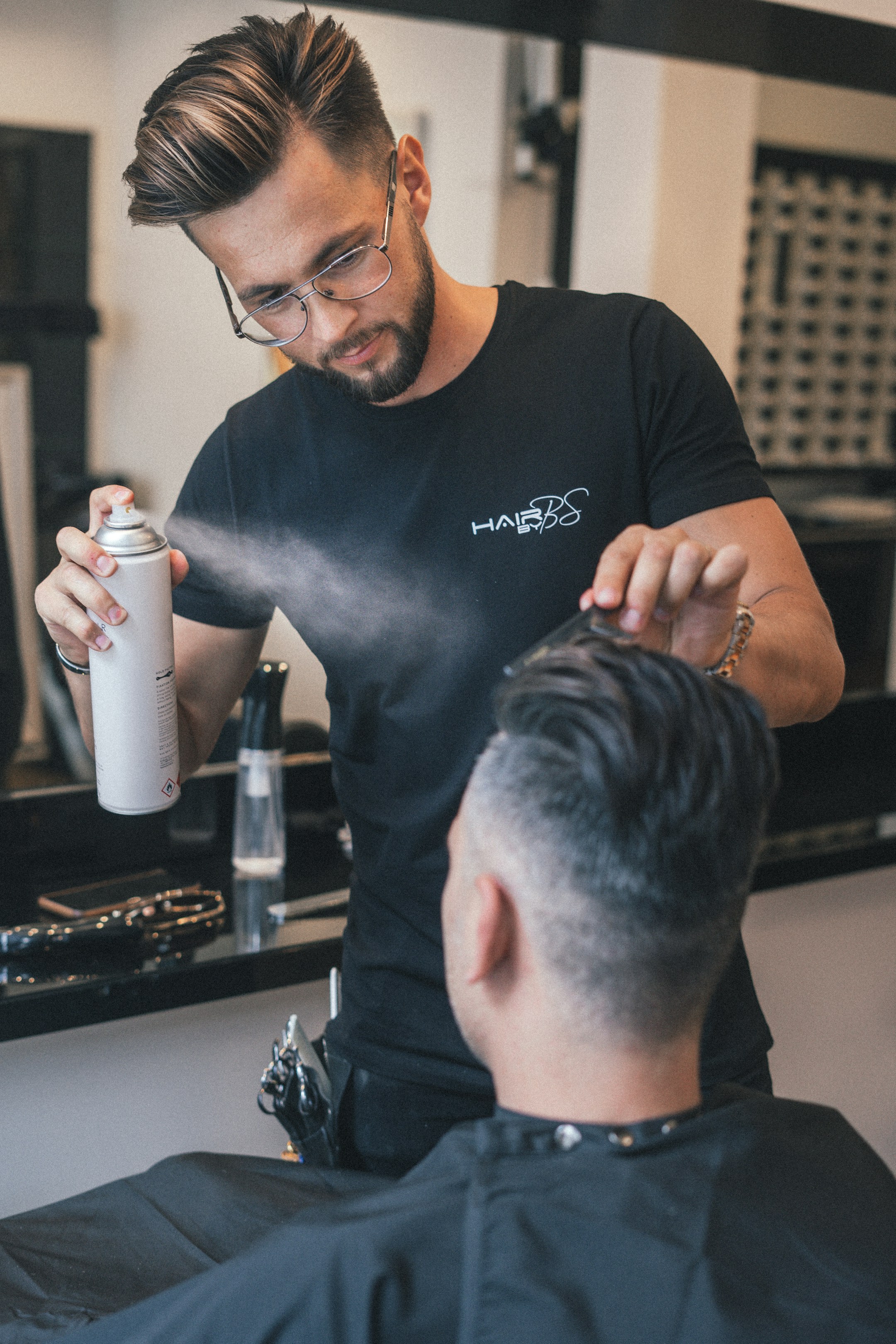 A barber portrait in a black shirt, holding a bottle of product, the calm authority of someone who runs a small shop.